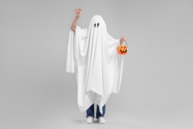 Woman in white ghost costume holding pumpkin bucket on light grey background. Halloween celebration