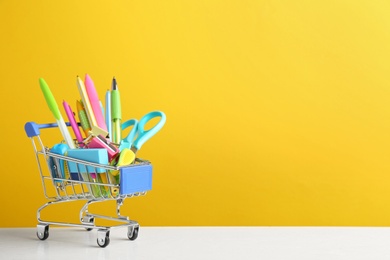 Small shopping cart with different school stationery on white wooden table against yellow background. Space for text