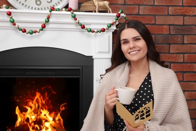 Photo of Young woman with greeting card and hot drink sitting near fireplace indoors