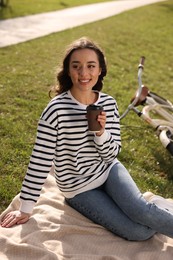 Young woman sitting on green grass and holding cup of coffee near bicycle outdoors