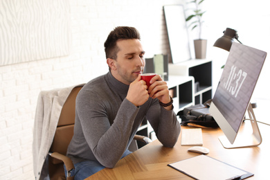 Young man with cup of drink relaxing at table in office during break