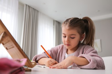 Adorable little girl doing homework with tablet at table indoors