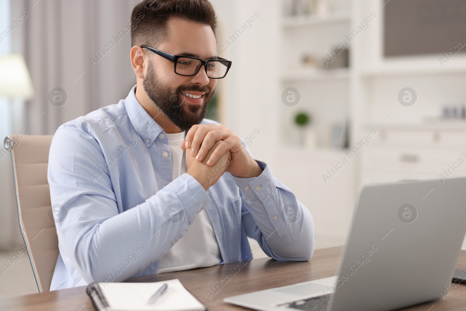 Photo of Young man in glasses watching webinar at table in room