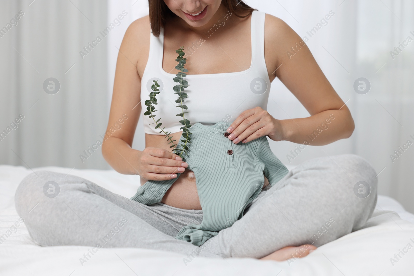 Photo of Pregnant woman with baby bodysuit and plant twigs sitting on bed indoors, closeup