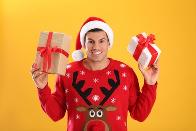 Handsome man in Santa hat holding gift boxes on yellow background