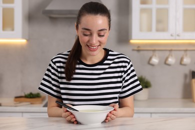 Photo of Smiling woman with bowl of tasty soup at white table in kitchen