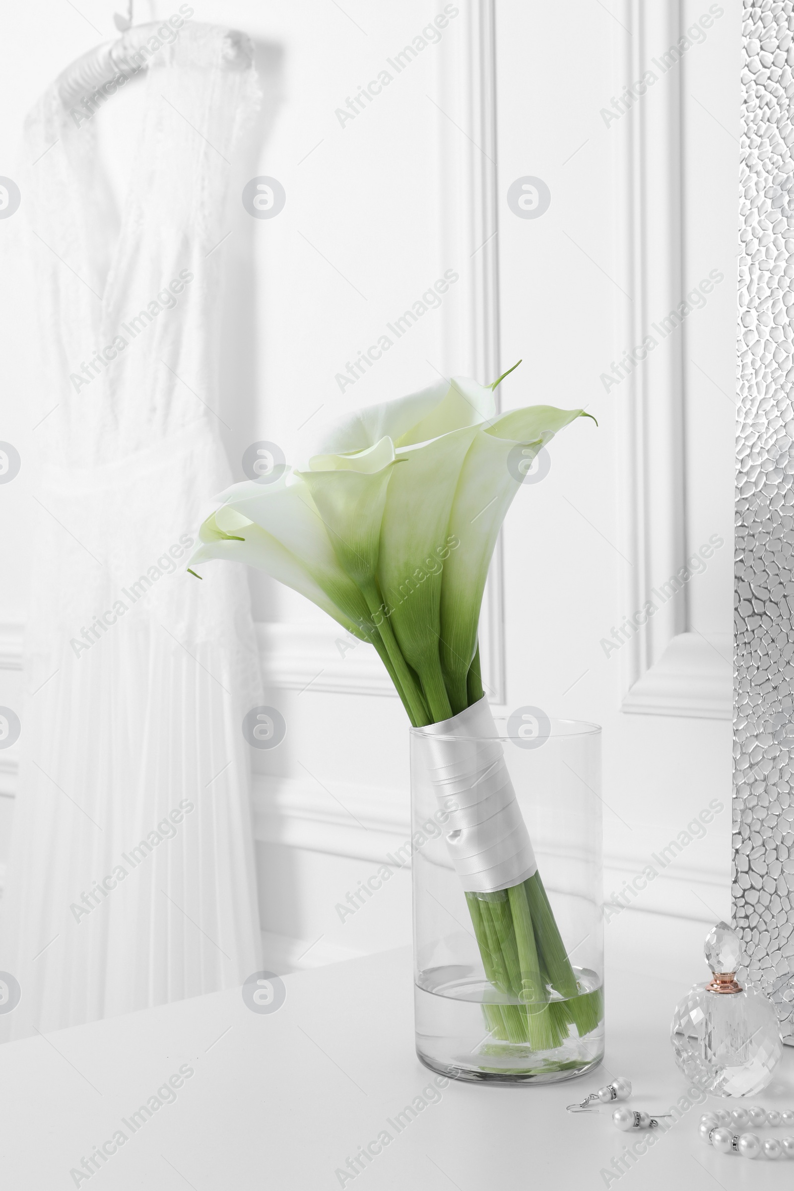 Photo of Beautiful calla lily flowers tied with ribbon in glass vase, bottle of perfume and jewelry on white table