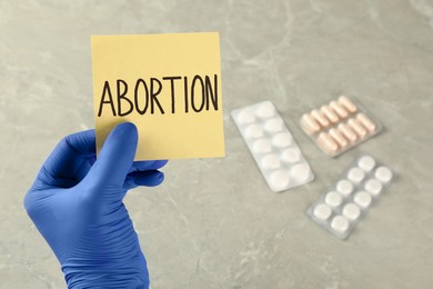 Photo of Doctor holding paper card with word ABORTION over light grey marble table with pills, closeup