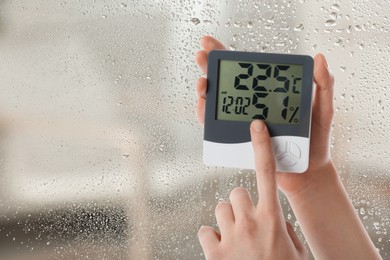 Woman holding digital hygrometer with thermometer near window on rainy day, closeup. Space for text