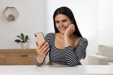 Photo of Happy young woman having video chat via smartphone at table in room