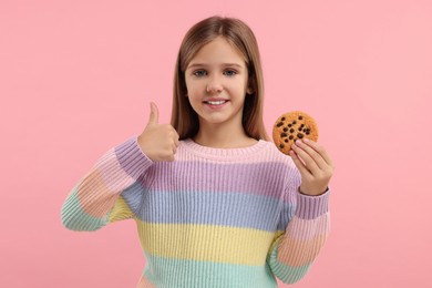 Photo of Cute girl with chocolate chip cookie showing thumb up on pink background