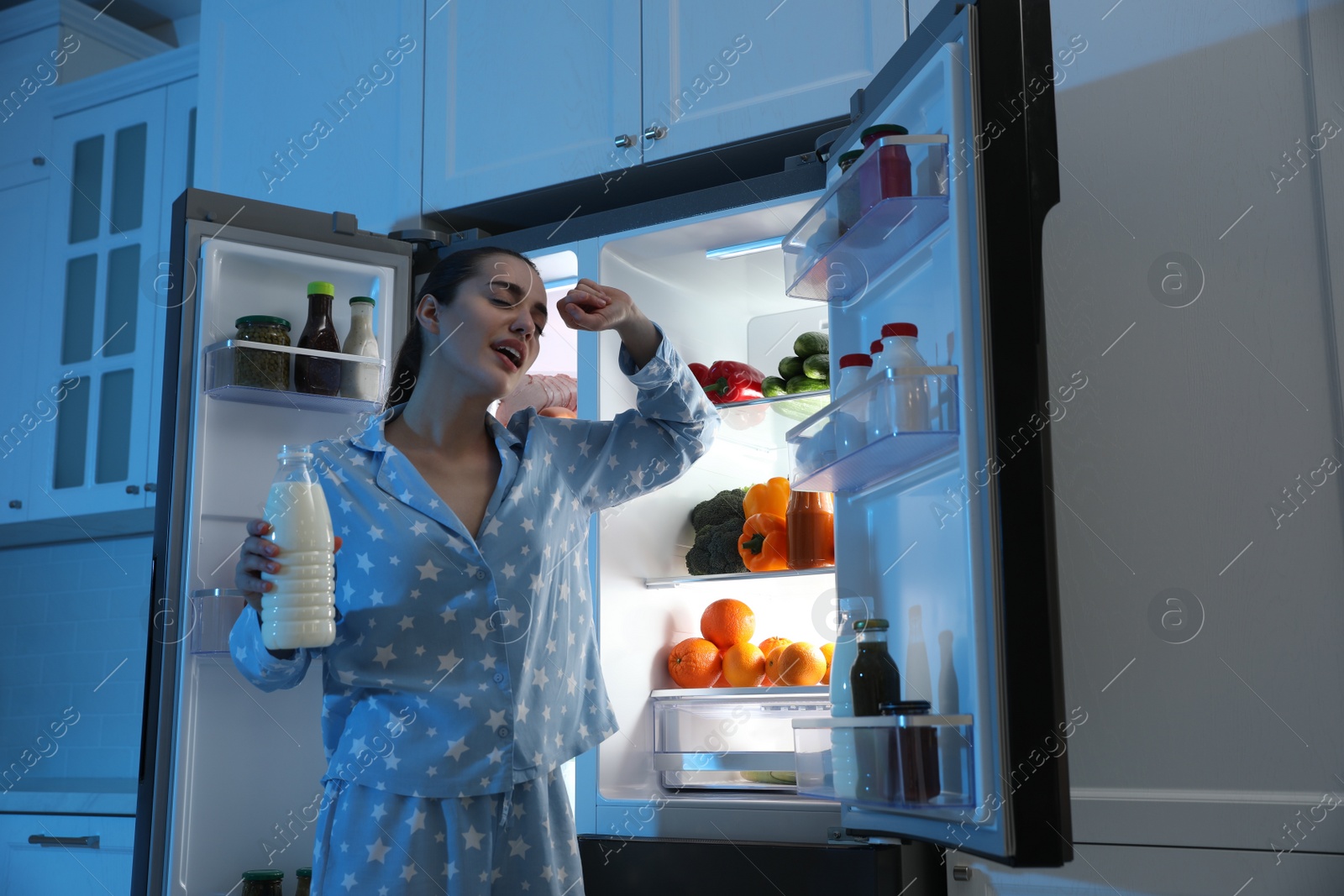 Photo of Young woman with bottle of milk near open refrigerator at night