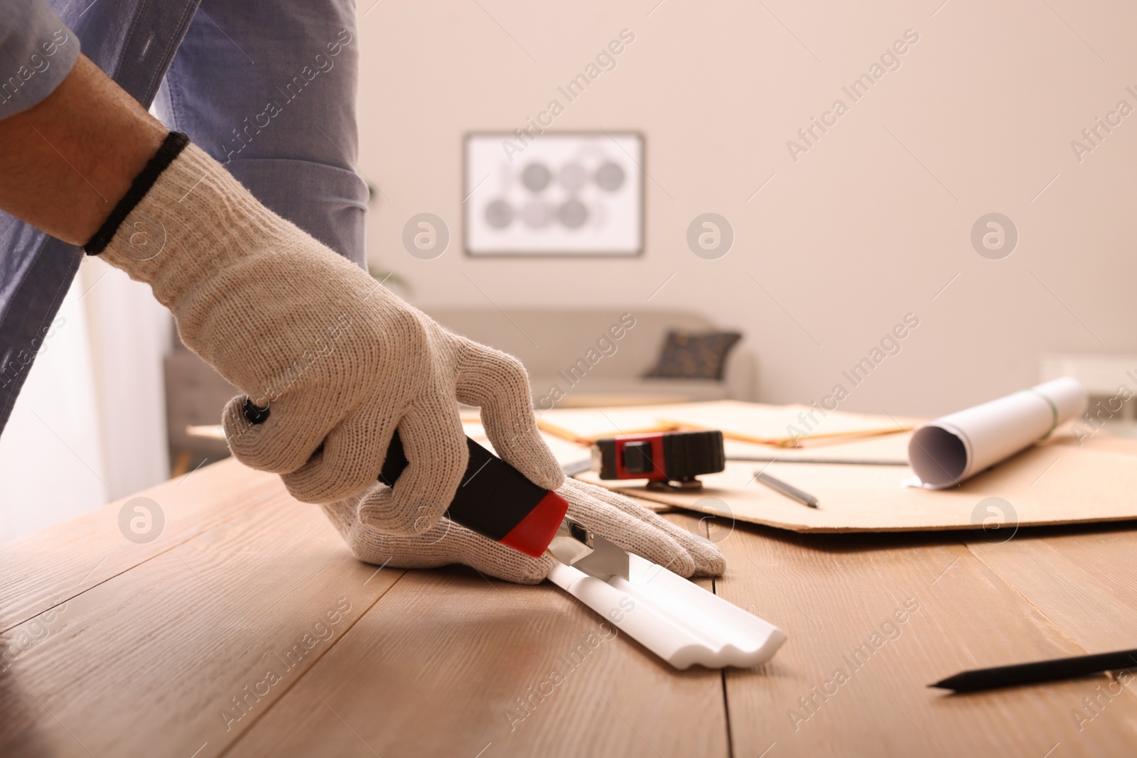 Photo of Man cutting foam crown molding with utility knife at wooden table indoors, closeup