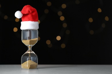 Photo of Hourglass with Santa hat on table against blurred lights, space for text. Christmas countdown