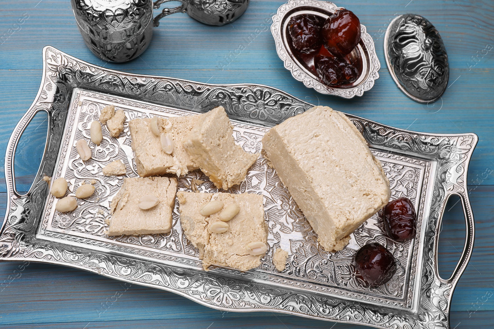 Photo of Pieces of tasty halva, dates and peanuts served in vintage tea set on light blue wooden table, above view