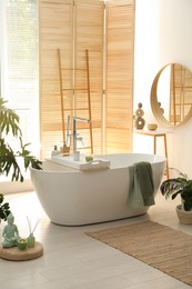 Stylish bathroom interior with modern tub, window and beautiful houseplants. Home design