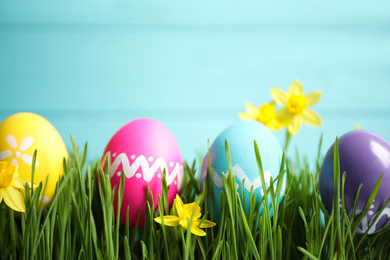 Photo of Colorful Easter eggs and narcissus flowers in green grass against light blue background