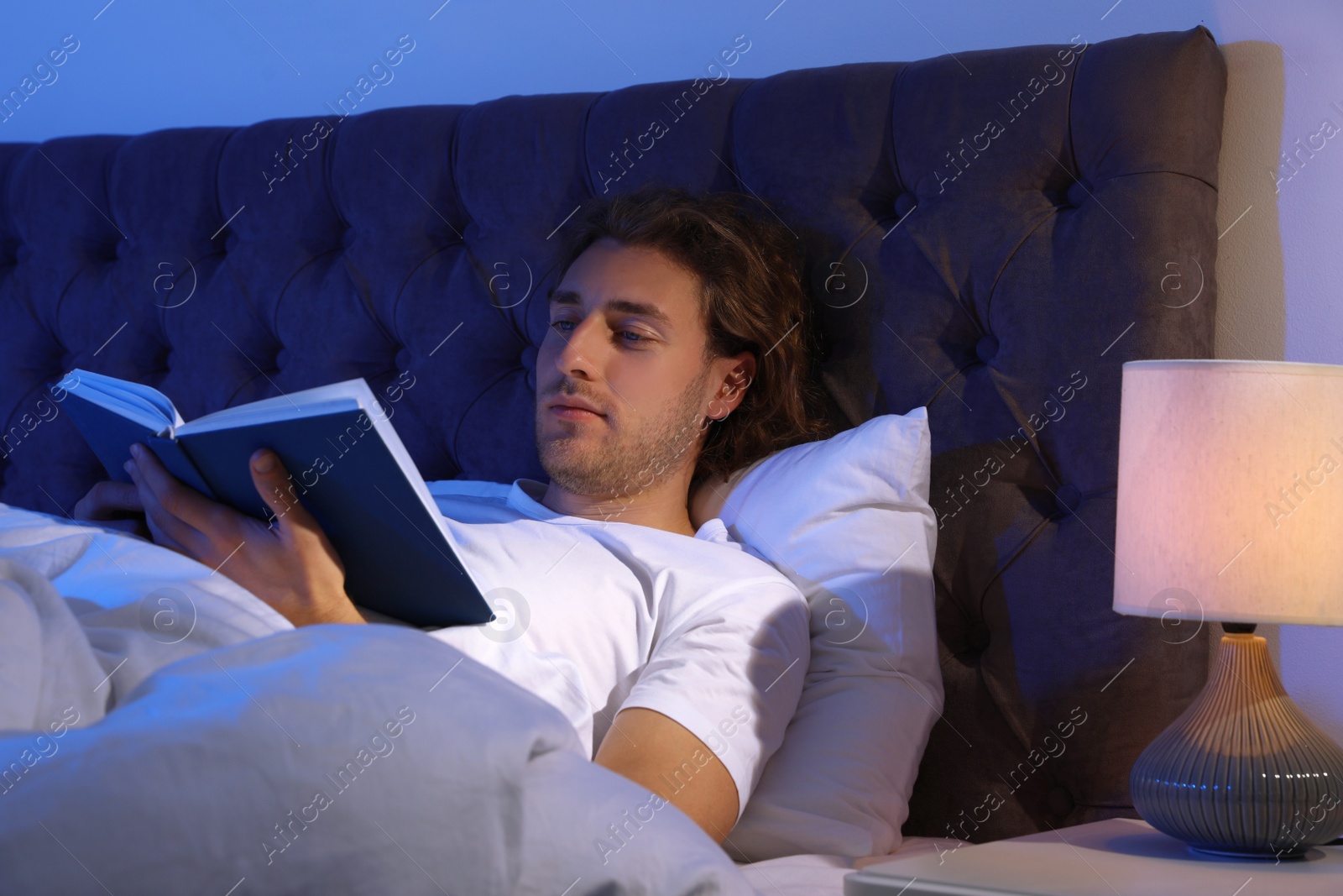 Photo of Handsome young man reading book in dark room at night. Bedtime