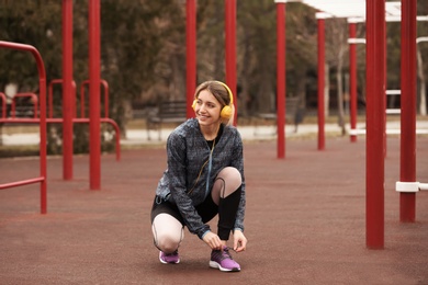 Photo of Young woman with headphones tying shoelaces on sports ground