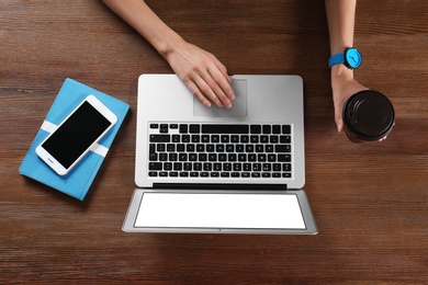 Photo of Young woman holding coffee while using laptop at wooden table, top view