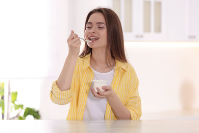 Photo of Young attractive woman eating tasty yogurt at table in kitchen