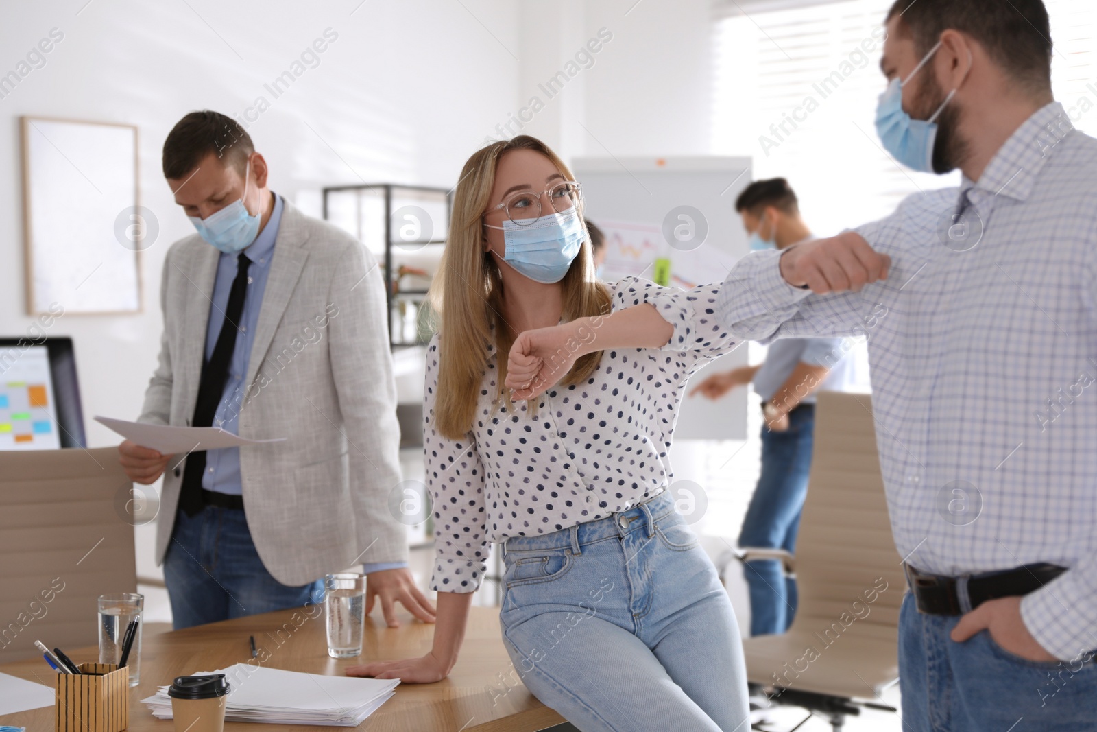 Photo of Coworkers with protective masks making elbow bump in office. Informal greeting during COVID-19 pandemic