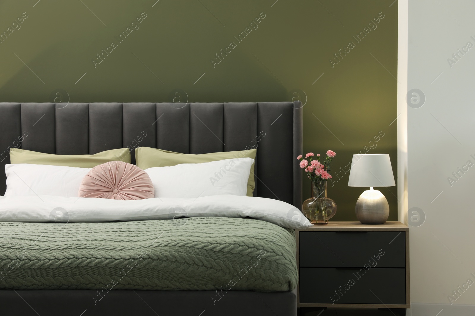 Photo of Large comfortable bed, nightstand, lamp and beautiful flowers in stylish room. Interior design