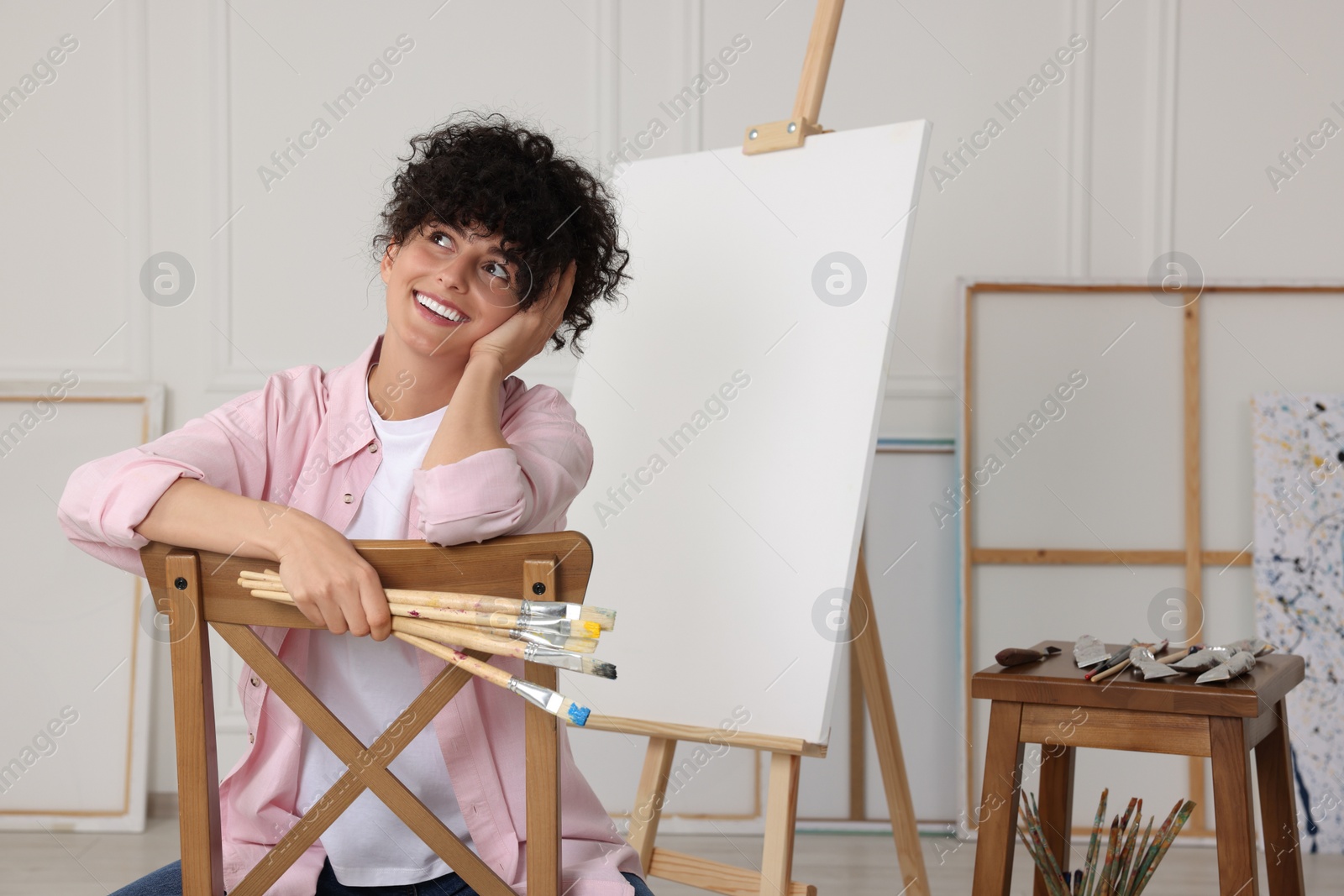 Photo of Young woman holding brushes near easel with canvas in studio