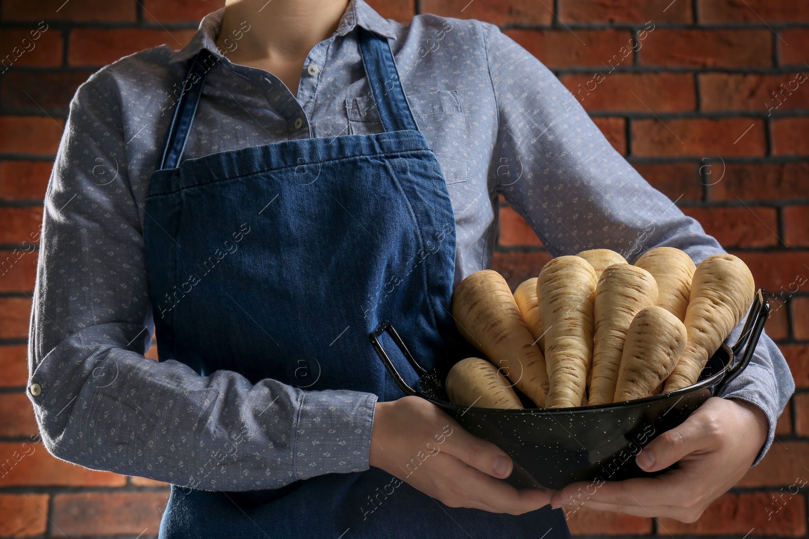 Photo of Woman holding bowl with fresh ripe parsnips near red brick wall, closeup