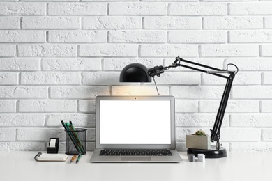 Photo of Modern workplace with laptop and lamp on table near brick wall. Space for design