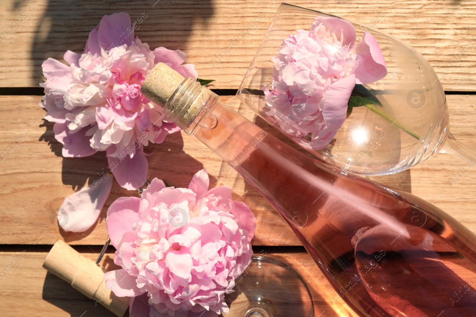 Photo of Bottle and glass of rose wine near beautiful peonies on wooden table, flat lay