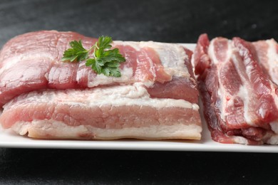 Photo of Pieces of raw pork belly and parsley on black textured table, closeup