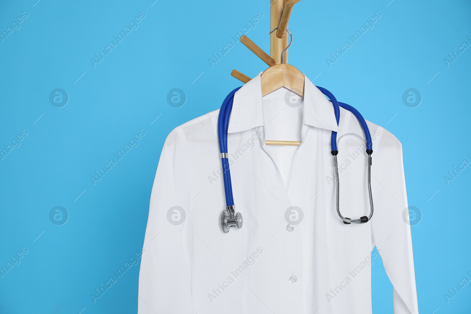 Photo of White medical uniform and stethoscope hanging on rack against light blue background. Space for text