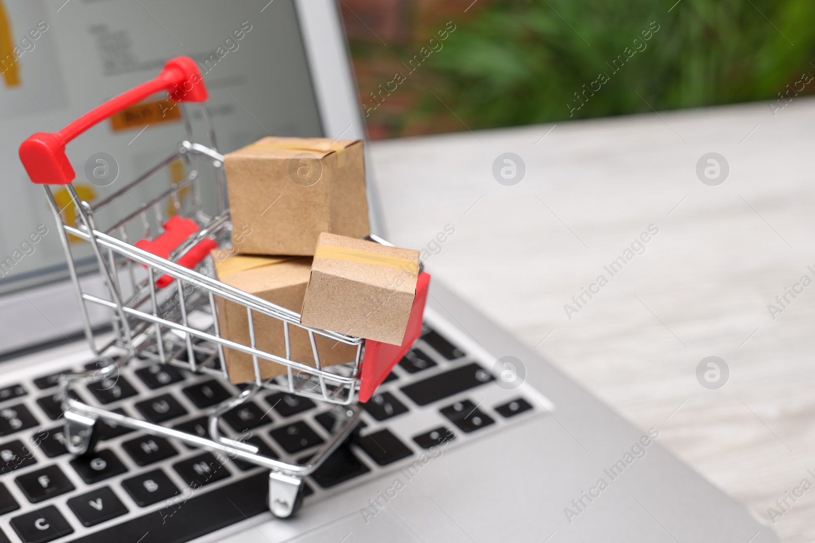 Photo of Internet store. Small cardboard boxes, shopping cart and laptop on light wooden table, closeup. Space for text