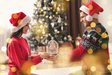 Couple in Santa hats holding snow globe in room. Bokeh effect