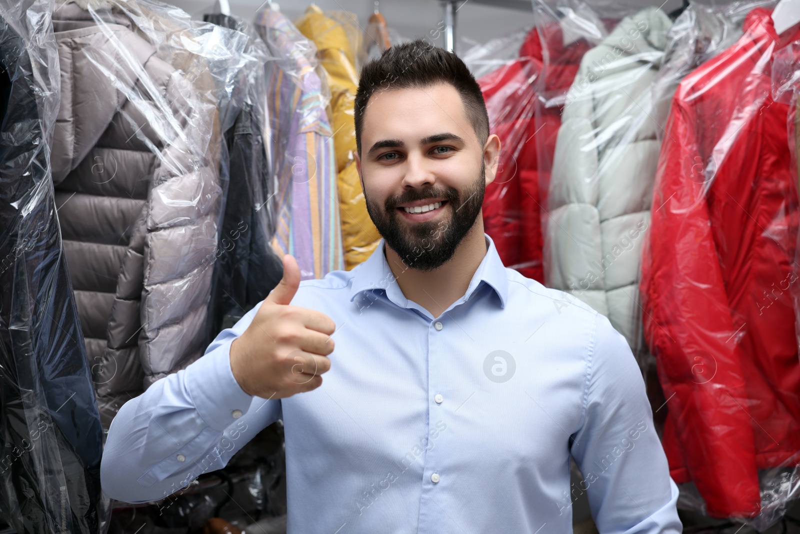 Photo of Dry-cleaning service. Happy worker showing thumb up near rack with different clothes indoors
