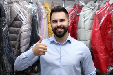 Photo of Dry-cleaning service. Happy worker showing thumb up near rack with different clothes indoors