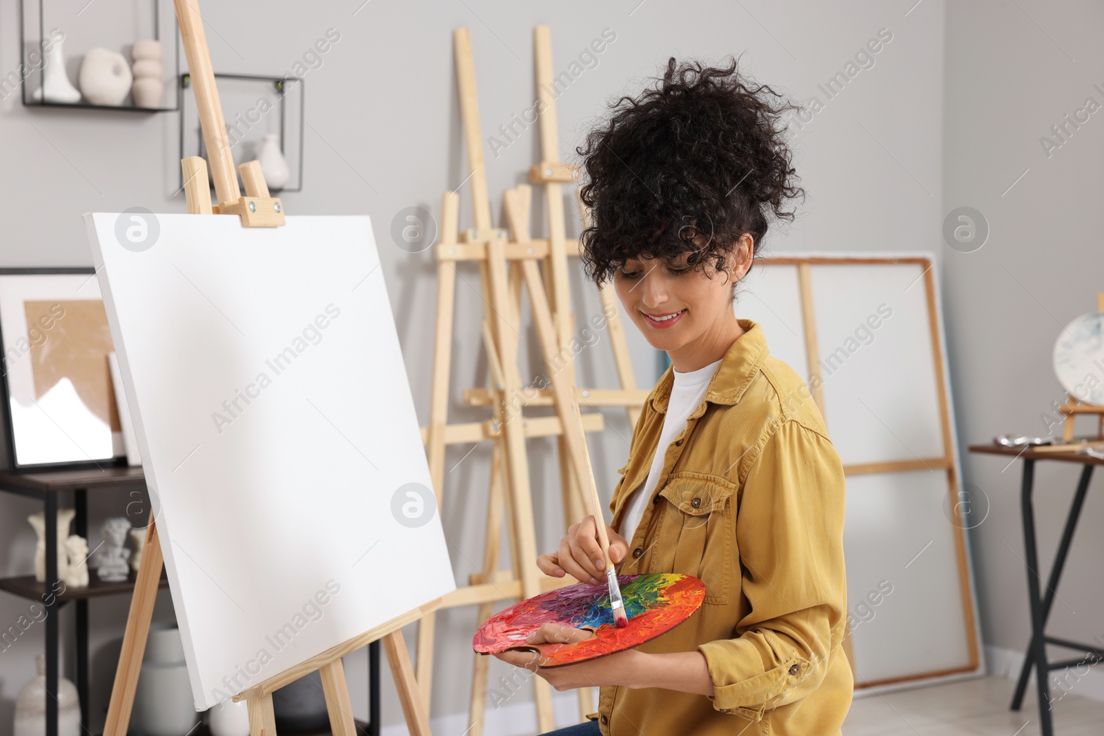 Photo of Young woman mixing paints on palette with brush near easel in studio