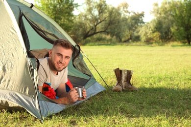 Photo of Young man in sleeping bag with cup of drink lying inside camping tent