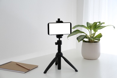 Smartphone with blank screen fixed to tripod on white table indoors. Mockup for design