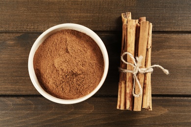 Aromatic cinnamon powder in bowl and sticks on wooden table, flat lay