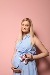 Photo of Beautiful pregnant woman in dress holding baby booties near tummy on color background