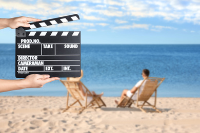 Assistant holding clapperboard and people relaxing in deck chairs on beach, closeup. Cinema production 