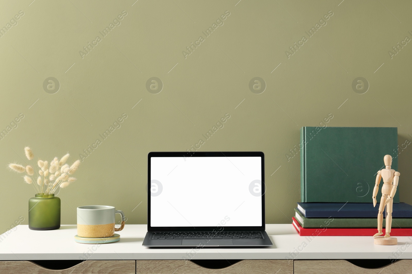 Photo of Stylish workplace with laptop, cup and vase of dry decorative spikes on white desk near olive wall indoors