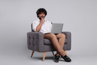 Young man with laptop sitting in armchair on light grey background