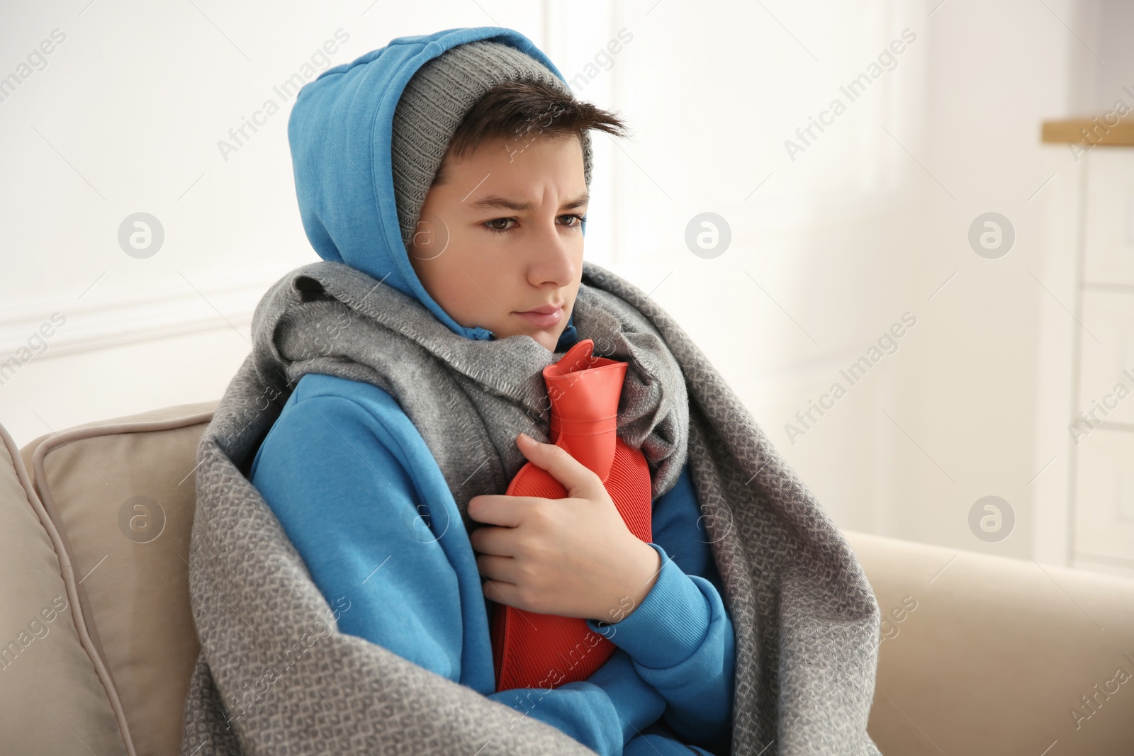 Photo of Sick teenage boy wrapped in blanket with hot water bottle on sofa at home