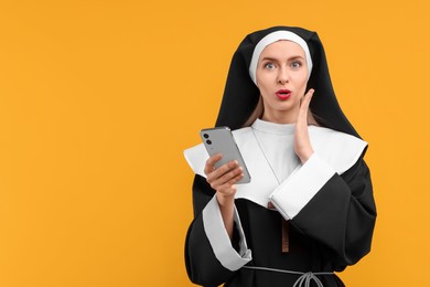 Photo of Surprised woman in nun habit with smartphone against orange background. Space for text