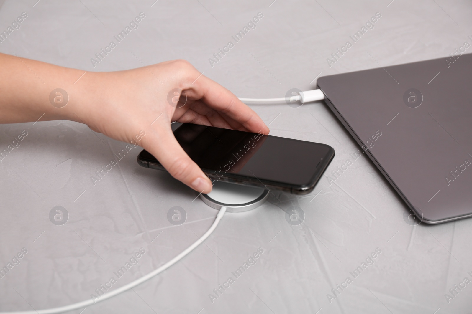 Photo of Woman putting smartphone on wireless charger connected to laptop at light grey table, closeup. Space for text