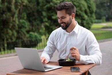 Photo of Happy businessman using laptop while having lunch at wooden table outdoors