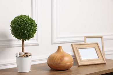 Green artificial plant in pot, frames and air humidifier on wooden table near white wall